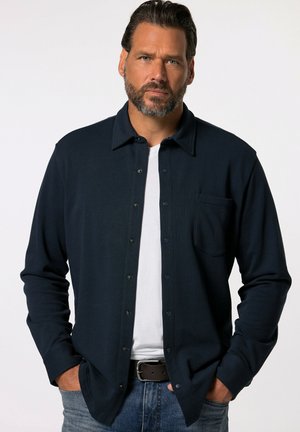 Camisa de manga larga azul marino con botonadura frontal, cuello y bolsillo. Presenta una textura acanalada y un corte relajado, combinada con una camiseta blanca de debajo.