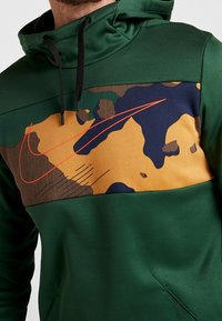 Homme portant un sweat à capuche vert foncé avec un panneau camouflage multicolore et un swoosh Nike orange sur la poitrine, cordons noirs visibles.