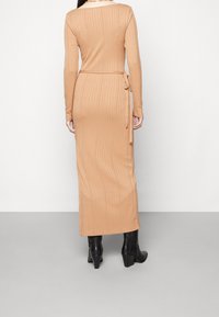 Robe longue côtelée en beige clair avec des manches longues et une ceinture à nouer à la taille. Modèle associé à des bottines noires à talons.
