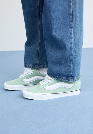 Henkilö, jolla on päällään vaaleanvihreät ja valkoiset sneakerit valkoisilla nauhoilla, sekä siniset farkut, seisoo vaaleansinisellä lattialla.