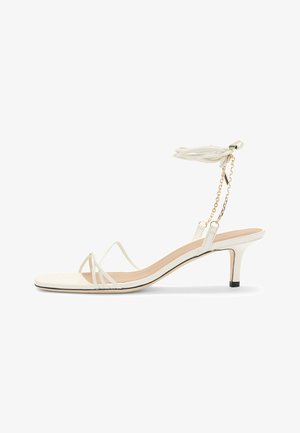 White strappy high-heeled sandal featuring thin straps, a square toe, a delicate ankle tie with tassel, and a gold-accented chain.