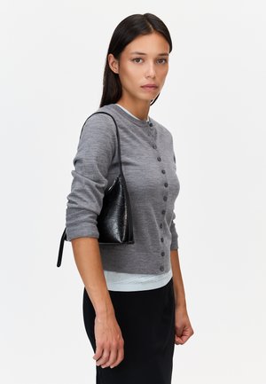 Femme aux cheveux lisses et foncés portant un pull gris boutonné et une jupe noire, portant un petit sac noir à l'épaule droite.