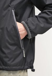 Czarna kurtka z kieszenią na zamek, o gładkiej fakturze i elastycznych mankietach. Charakterystyczne detale to sznurek ściągający u dołu oraz suwak z logo.