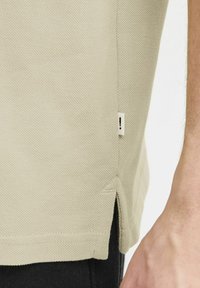 Lys beige polo-skjorte med teksturert stoff. Har sidesplitter ved kanten og et lite stoffmerke på siden.