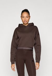 Καφέ cropped φούτερ με κουκούλα, άνετη εφαρμογή και μακριά μανίκια. Διαθέτει οριζόντια ρίγα στο κάτω μέρος. Συνδυάζεται με κολάν στο ίδιο χρώμα.