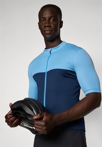 Maillot de cyclisme en bleu clair et marine, à manches courtes, fermeture éclair, texture lisse. Le modèle tient un casque noir, avec un arrière-plan simple.