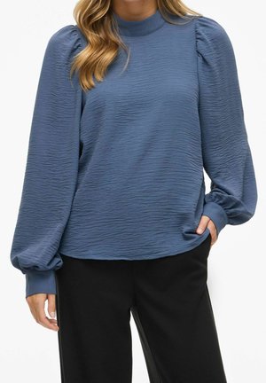 Kobieta ubrana w niebieską bluzkę z długim rękawem o fakturze i bufiaste rękawy oraz czarne spodnie, swobodnie trzymająca rękę w kieszeni.