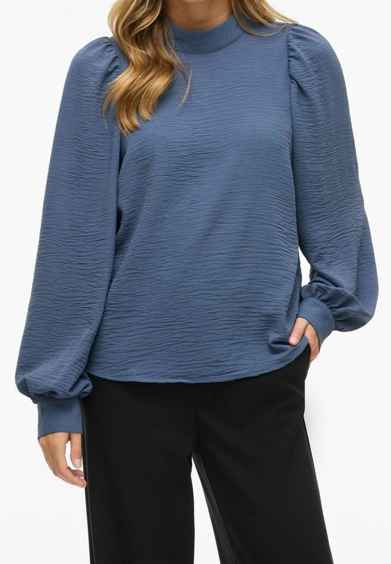 Femme portant un chemisier bleu texturé à manches longues bouffantes et un pantalon noir, la main posée de manière décontractée dans la poche.