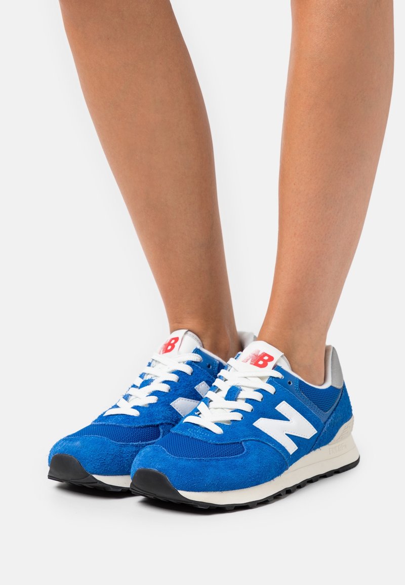 Blå sneakers i mocka och mesh, med vita accenter och snören, en svart gummisula och röd branding på tungan.