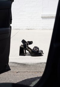 Paire de sandales à talons hauts en cuir verni noir avec boucles, posées sur un bord de trottoir, vue depuis la portière ouverte d'une voiture.