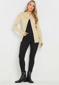 Gilet en tricot beige à col rond, avec une fermeture à boutons sur le devant et des manches longues, associé à un jeans noir ajusté et des bottines noires.