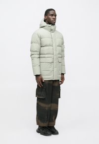 Mand står sidelæns iført en lysegrøn hættetrøje i puffer-stil, sorte og brune brede cargo bukser og sorte sneakers foran en hvid baggrund.