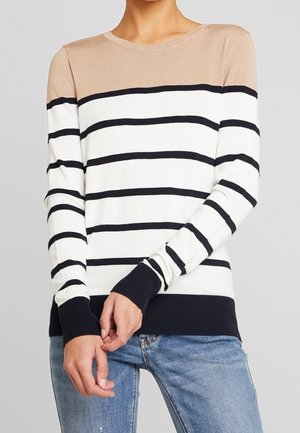 Femme portant un pull à manches longues rayé beige, blanc et noir avec poignets et ourlet côtelés, associé à un jean bleu, les mains jointes devant.