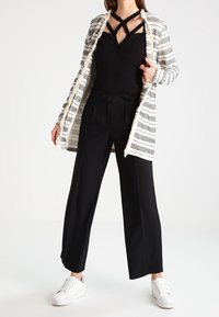 Kobieta ubrana w czarny top na ramiączkach, wzorzysty długi kardigan, czarne szerokie spodnie i białe trampki, stojąca z rozstawionymi nogami.