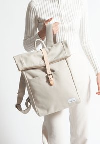 Beige canvasryggsäck med ett överliggande lock, dragkedjestängning, läderrem och framficka. Strukturerad tyg och justerbara remmar.