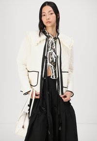 Κρεμ σακάκι με καπιτονέ σχέδιο, διακοσμημένο με ριχτές κολάρο και μαύρες λεπτομέρειες, φορεμένο πάνω από ένα λουλουδάτο μπλουζάκι. Το μοντέλο κρατά μια λευκή τσάντα. Μαύρη φούστα με κορδέλες.