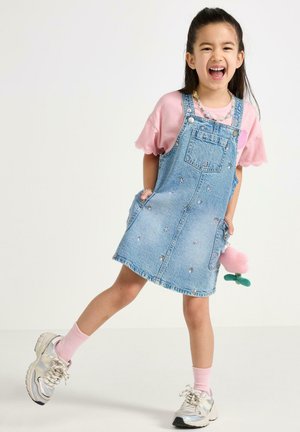 Glunderend meisje met een roze shirt en een spijkerjurk met borduursels, roze sokken en sneakers, poserend met een opgetrokken been.