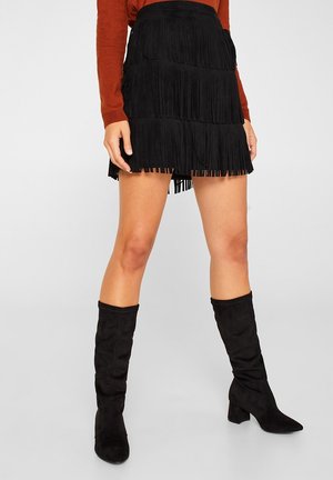 Femme portant une mini-jupe noire à étages avec franges, des bottes noires montantes et un haut à manches longues couleur rouille, debout devant un fond clair uni.