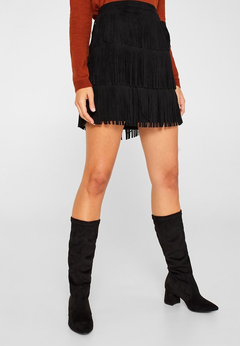 Femme portant une mini-jupe noire à étages avec franges, des bottes noires montantes et un haut à manches longues couleur rouille, debout devant un fond clair uni.