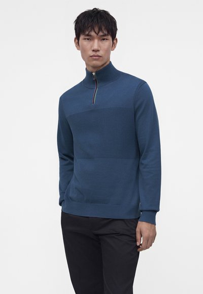 Jeune homme portant un pull bleu texturé à demi-fermeture éclair et un pantalon noir, debout devant un fond blanc uni.