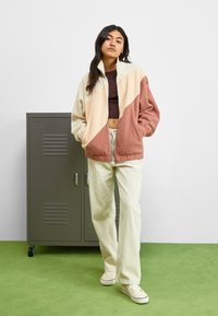 Chaqueta de forro polar en bloques de color crema, rosa y beige con cuello alto, combinada con pantalones de pana claros y zapatillas blancas.