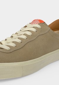 Baskets en daim beige avec lacets blancs, semelle beige clair et détail du logo orange sur la languette, sur un fond uni.