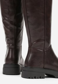Bottes en cuir marron jusqu'au genou avec un design épuré, dotées d'une semelle en caoutchouc noir épaisse et de détails de couture subtils.