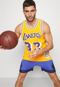 Maglietta da basket gialla in rete con accenti viola, con la scritta "Lakers" e il numero "32". Accompagnata da pantaloni blu con strisce nere e un pallone da basket.