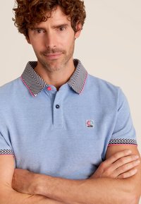 Lichtblauwe polo met een zwart-wit gestreepte kraag, accentueert met een roze piping. Bevat een klein logo op het linkerbovenborstgebied.