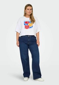 Balta īsām piedurknēm t-kreklu ar krāsainu tomātu grafiku, apvienota ar brīvā griezuma tumši ziliem džinsiem un baltiem sporta apaviem. Vienkāršs, ikdienišķs apģērbs.