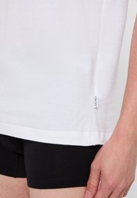 Vit bomull T-shirt med kort ärm, med en sidolapp. Buren över svarta tygshorts, som visar en slät textur och enkel design.