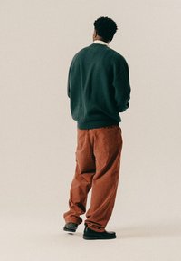 Dark green oversized sweatshirt, orange corduroy pants with a checkered pattern, black shoes, viewed from the back against a neutral background.