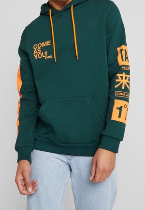 Mann trägt dunkelgrünen Hoodie mit orangem Schriftzug und Kordeln, kombiniert mit hellblauen Jeans, steht mit entspannten Händen.
