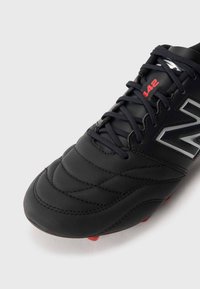 Black football shoe with quilted texture, lace closure, silver logo, and red accents on the outsole. Features round red studs for traction.