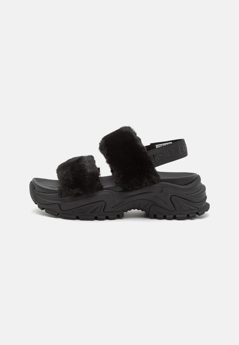 Kraftig sort sandal med tyk, tekstureret sål, to brede sorte faux pels-remme og en justerbar branded bagrem.