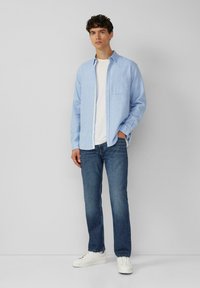 Lichtblauw button-up shirt met een voorzak, witte t-shirt eronder, gecombineerd met donkerblauwe jeans en witte sneakers. Effen achtergrond.