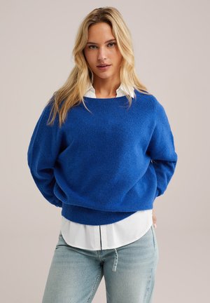 Blauwe wollen mix trui met een ontspannen pasvorm, brede halslijn en geribbelde zoom, gedragen over een wit shirt. Zachte textuur, casual ontwerp.