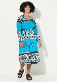 Světle modré šaty s černobílými vzory, s výstřihem do V, dlouhými rukávy a volným střihem. Černé sandály doplňují outfit.