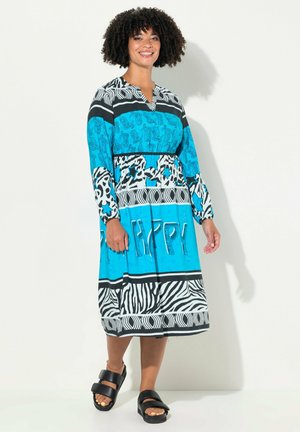Lichtblauwe jurk met zwart-witte patronen, met een V-hals, lange mouwen en een losse pasvorm. Zwarte sandalen maken de outfit compleet.