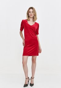 Rotes Samtkleid mit kurzen Puffärmeln, V-Ausschnitt und figurnahem Design. Beinhaltet ein kleines dekoratives Detail nahe dem Saum. Kombiniert mit schwarzen High Heels.