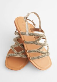 Sandali piatti beige con cinturini metallici intrecciati e fibbia regolabile alla caviglia, dotati di una suola liscia e texturizzata e ritagli geometrici.