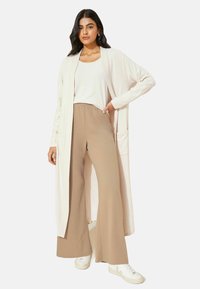 Lys beige lang cardigan med åben front, to lommer og ribbet tekstur. Sammen med beige vide bukser og hvide sneakers.