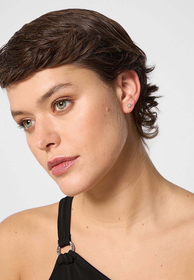 Orecchini a perno in tono oro con design floreale, indossati da una persona. Gli orecchini presentano dettagli intricati e una finitura lucida.