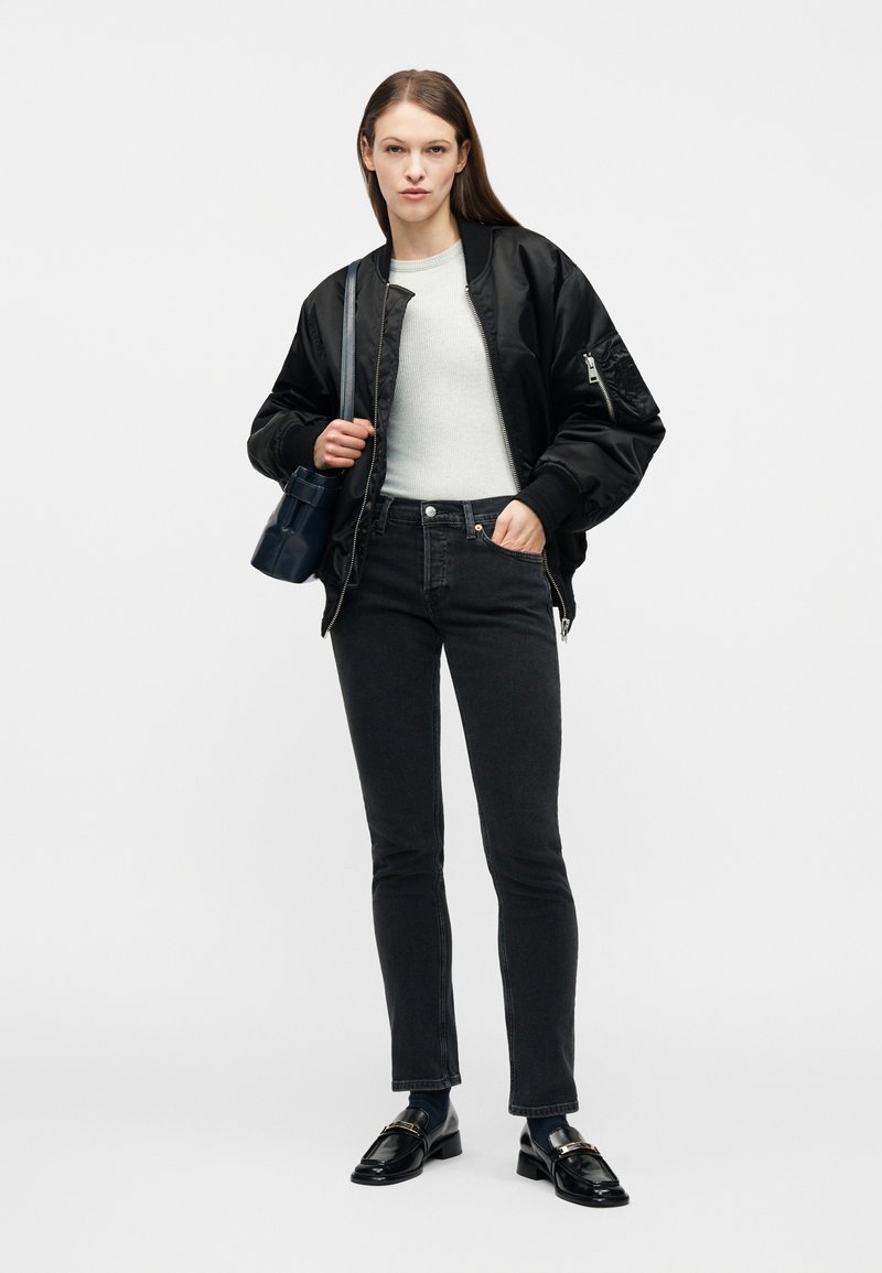 Young woman wearing black bomber jacket, white sweater, black jeans, black loafers, and carrying a shoulder bag, standing against white background.