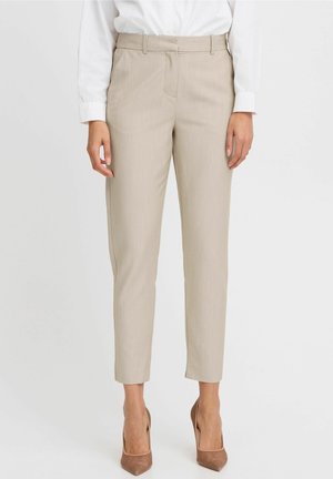 Frau trägt beigefarbene, maßgeschneiderte, knöchellange Hosen, weißes, eingestecktes Hemd und braune, spitz zulaufende High Heels, steht vor einem schlichten Hintergrund.