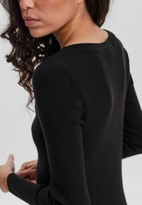 Haut noir à manches longues côtelé avec une coupe ajustée. Présente un col rond et une texture subtile. Les cheveux bouclés du modèle sont visibles.