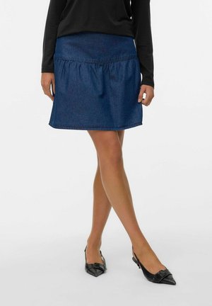Vrouw draagt een donkerblauwe denim rok, zwarte top met lange mouwen en zwarte pumps met spits toelopende neus en slingback, staand met gekruiste benen.