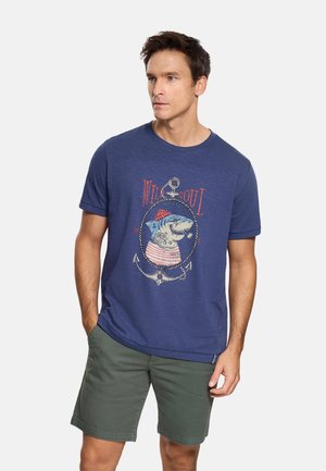 Hombre con camiseta azul con un gráfico náutico de tiburón y pantalones cortos verdes, de pie con una mano en el bolsillo sobre un fondo liso.