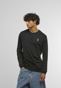 Melna garā piedurkne džemperis ar apaļu kaklu, kuram uz kreisā krūšu ir neliels balts logotipa akcenta. Izgatavots no kokvilnas materiāla, brīvs piegriezums.