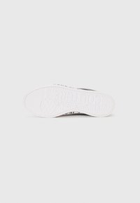 Hvit sneaker med en teksturert gummisåle. Svart merkevare på siden, minimalistisk design og en jevn kanvasoverdel. Toppvinkel.